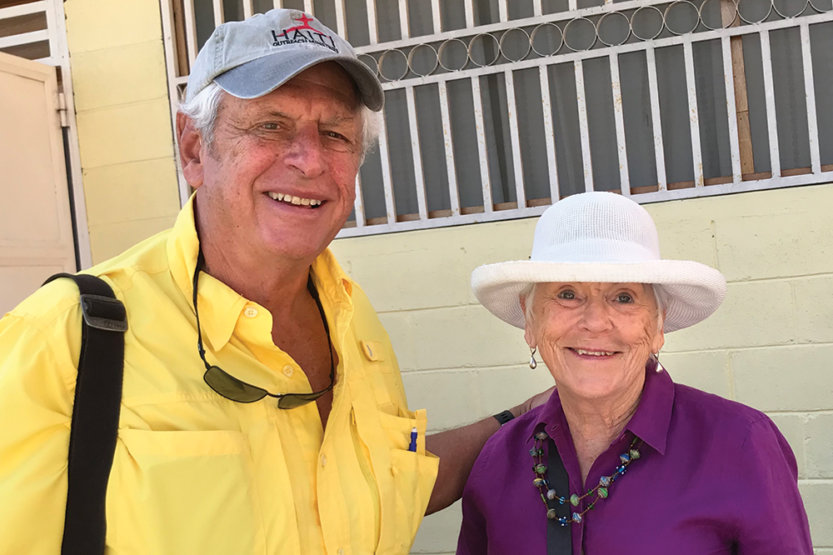 Bob and Flo Anne O’Brien in front of yellow painter cinderblock wall in Haiti.