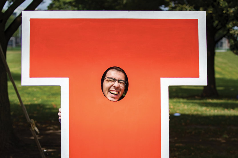 Male student being silly, laughing and poking his face through a life-size, orange Illinois block "I."