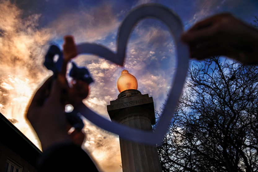 Dusk shot of the eternal flame, which is a light atop a stone column, through a heart held by hands.