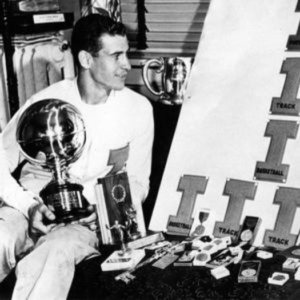 Dike Eddleman in letterman sweater seated on a table among his many awards and trophies.
