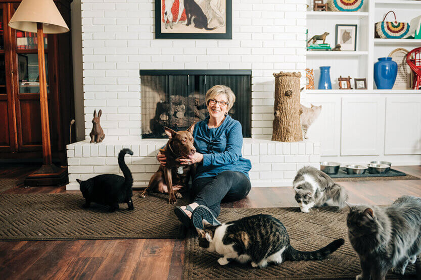 Gayle Magnuson seated on a fireplace hearth in her home surrounded by dogs and cats.