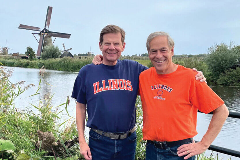 Larry Weber and Wes Jaros, wearing Illini t-shirts pose by a river in front of an old windmill.