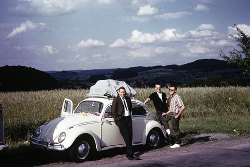Three young men stand, on a country road, around a white Volkswagen Beetle.