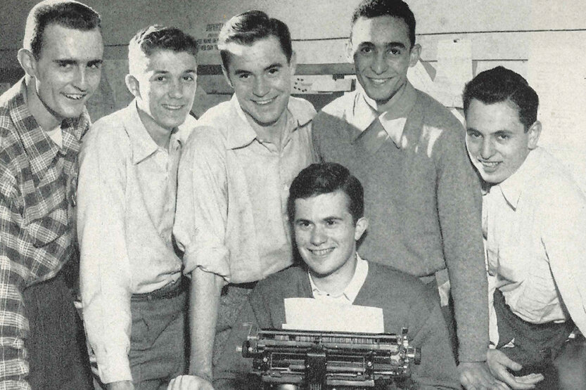 Morris Beschloss seated in front of a typewriter with 5 male colleages standing behind him.
