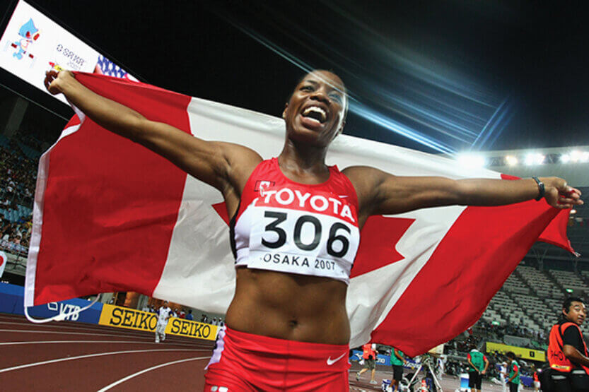 Perdita Felicien on the track celebrates her 100m Hurdles second-place finish holding a Canadian flag above her head.