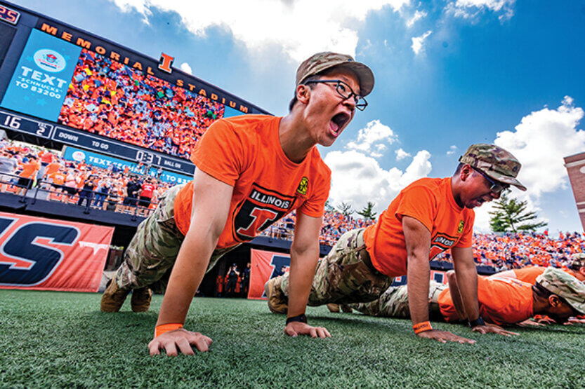 Members of the Illini ROTC do pushups on the football field in a packed stadium.