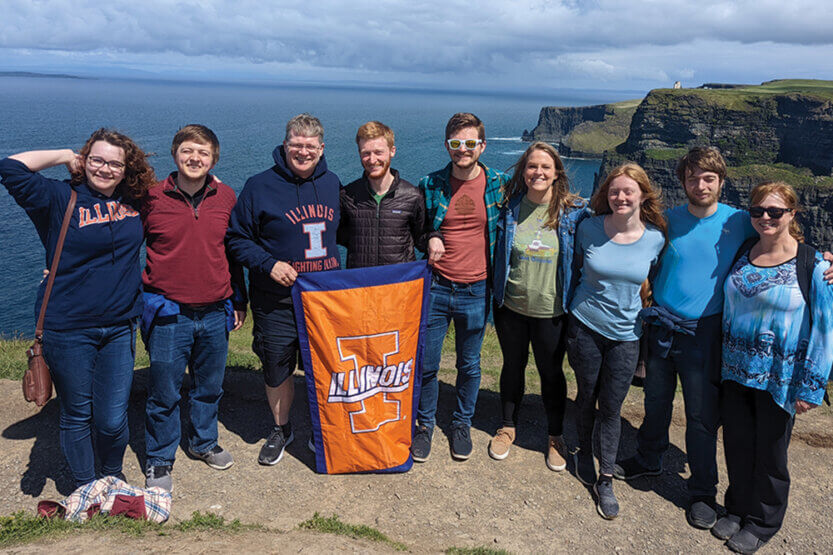 Individuals on a cliff overlooking the ocean holding an orange Illinois flag