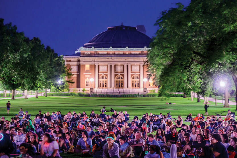 Students seated outside Illini Union at dusk watching projected film.