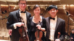 Sam Walder with two other musicians holding violins on stage at a concert hall