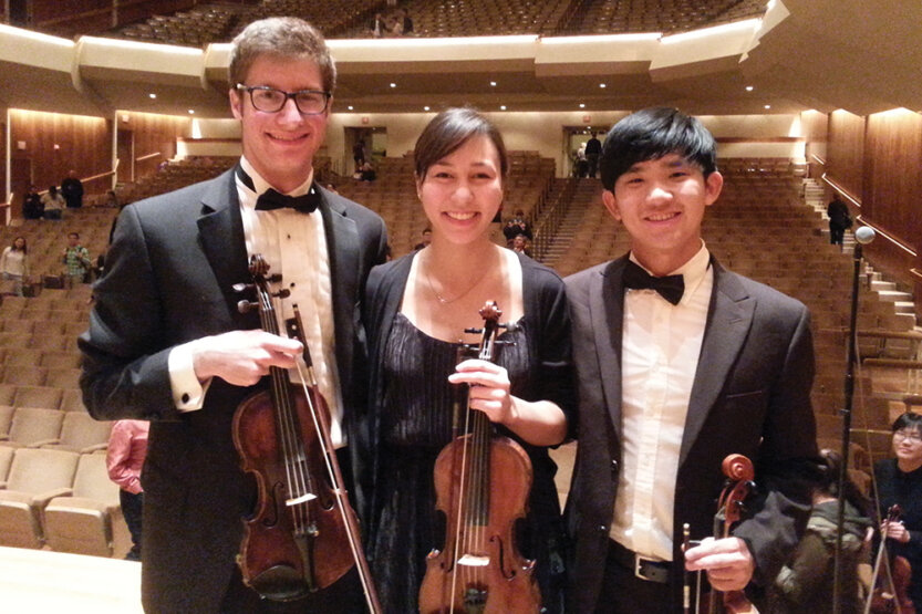 Sam Walder with two other musicians holding violins on stage at a concert hall